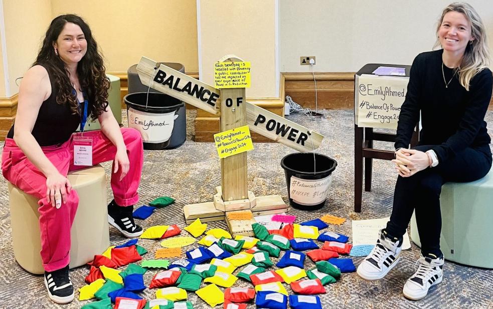 Emily and her colleague sitting on stools either side of Emily's 'balance of power' wooden scales, surrounded by colourful beanbags with labels attached.